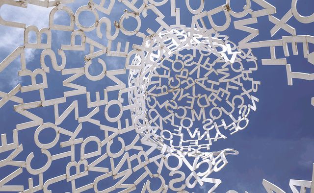 Looking up through a sculpture of metal letters spiralling up towards a blue sky.