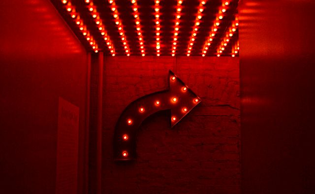 Looking down the end of a red lit hallway. A metal wall sculpture of red light bulbs inside an arrow pointing right.